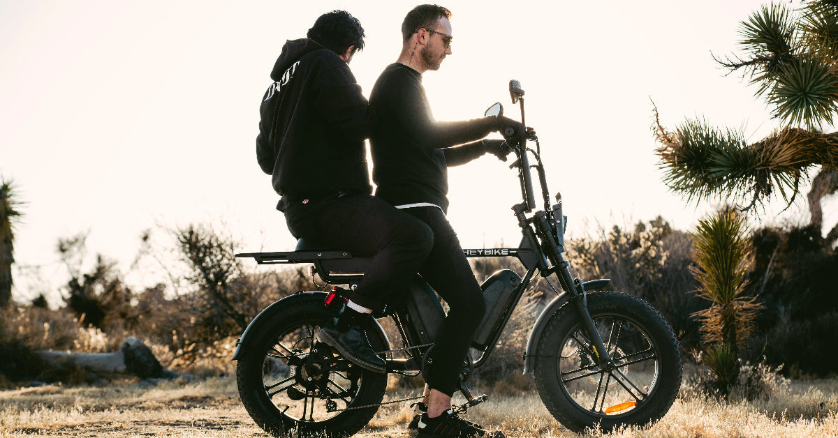 Two people riding a Heybike Saturn e-bike on a dusty trail with a desert landscape in the background.