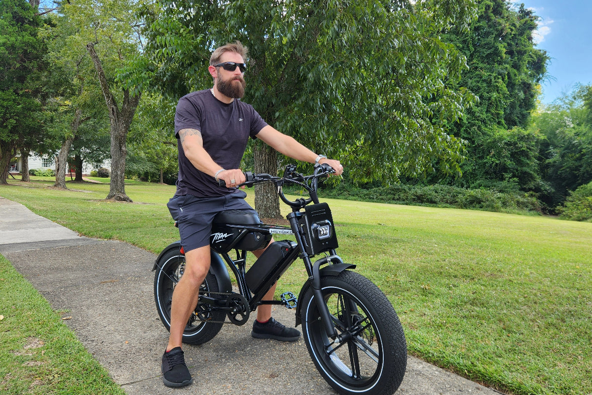 A man wearing sunglasses rides a black Heybike Titan electric bike on a paved path in a green park.