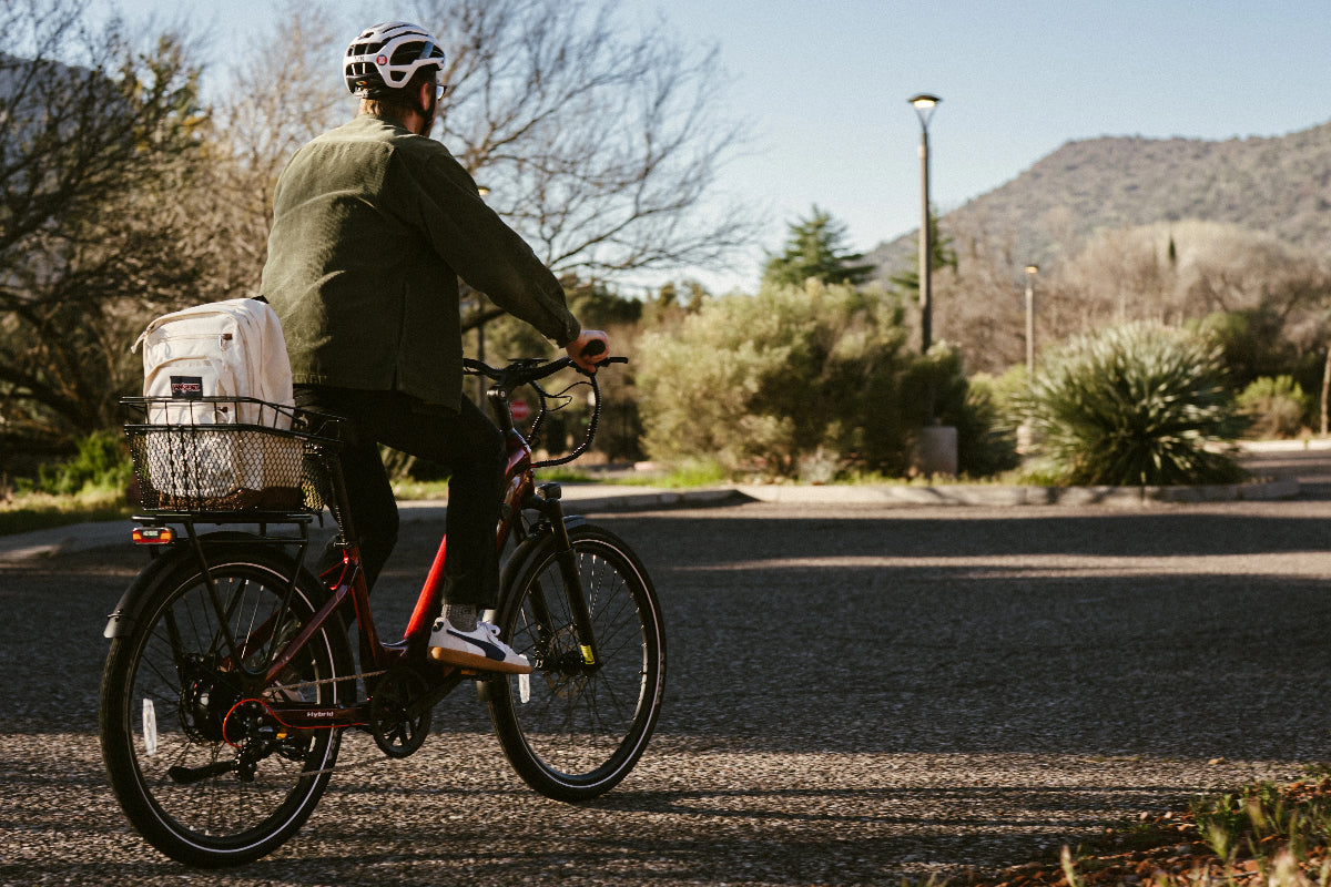 A cyclist wearing a helmet rides a black step-through electric bike with a rear basket along a quiet road with mountains in the background.